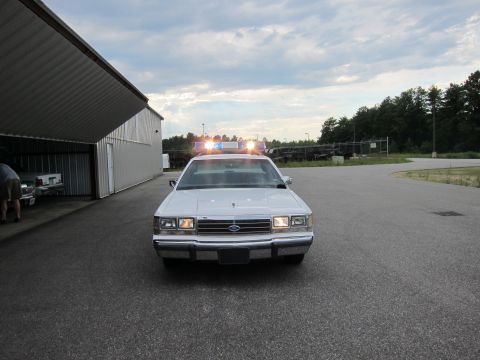 1991 Ford LTD Crown Victoria - A white police car sits in front of a large open-sided building with its emergency lights illuminated.