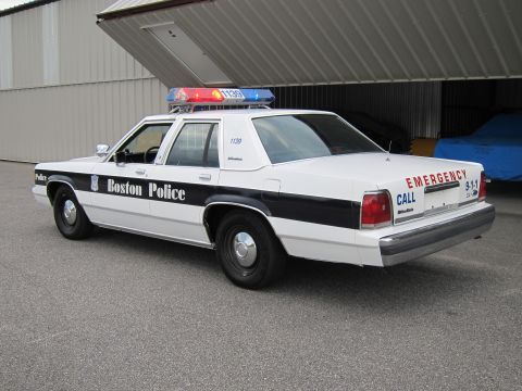 1991 Ford LTD Crown Victoria - A white police car with a light bar is parked in front of a large open garage door.
