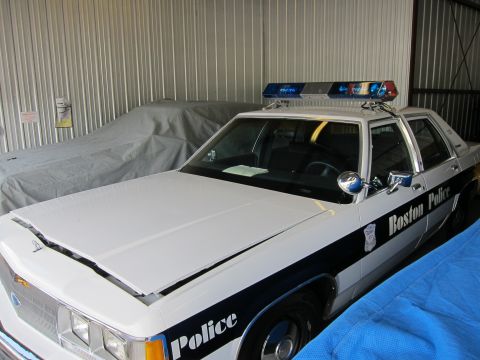 1991 Ford LTD Crown Victoria - A white 1991 Ford Crown Victoria LTD police car with "Boston Police" text is parked in a garage next to a covered vehicle.