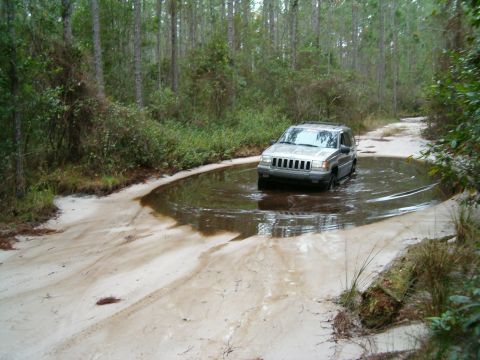 1991 Ford Bronco - A silver Jeep Grand Cherokee navigates a muddy puddle on a dirt road surrounded by trees.