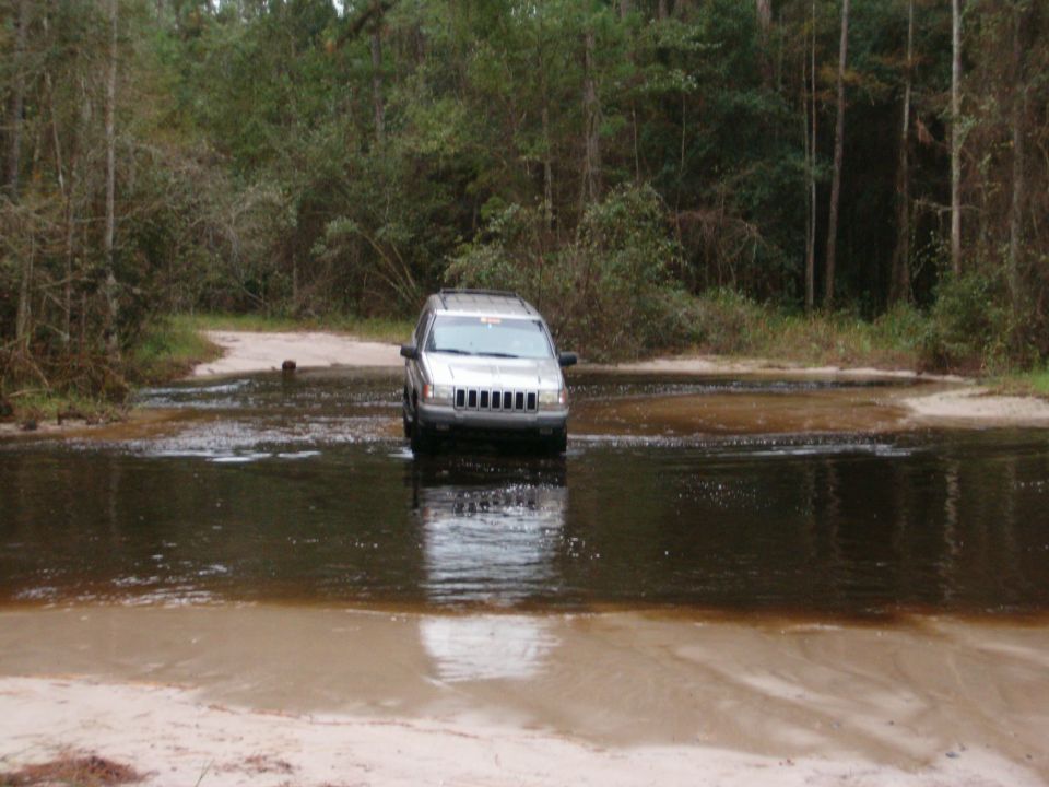 Silver Jeep Grand Cherokee fording a flooded sandy forest road, water up to the rocker panels