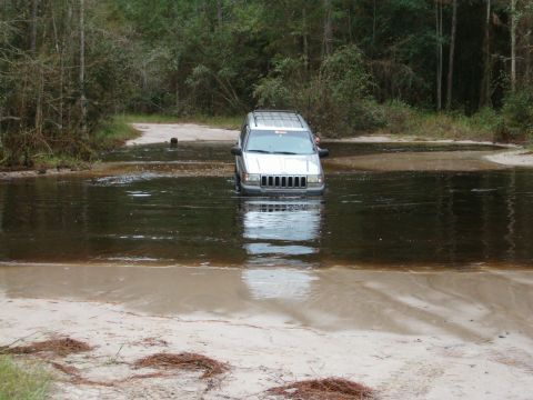 1991 Ford Bronco - Silver Jeep Grand Cherokee navigates a water crossing in a wooded area.
