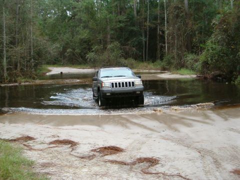 1991 Ford Bronco - A Jeep Grand Cherokee navigates a water crossing in a forest setting.