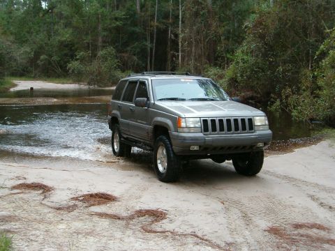 1991 Ford Bronco - A gray Jeep Grand Cherokee ZJ Laredo with roof rack, CB whip antenna and mud-terrain tires on alloy wheels pulls out of a flooded sand trai…