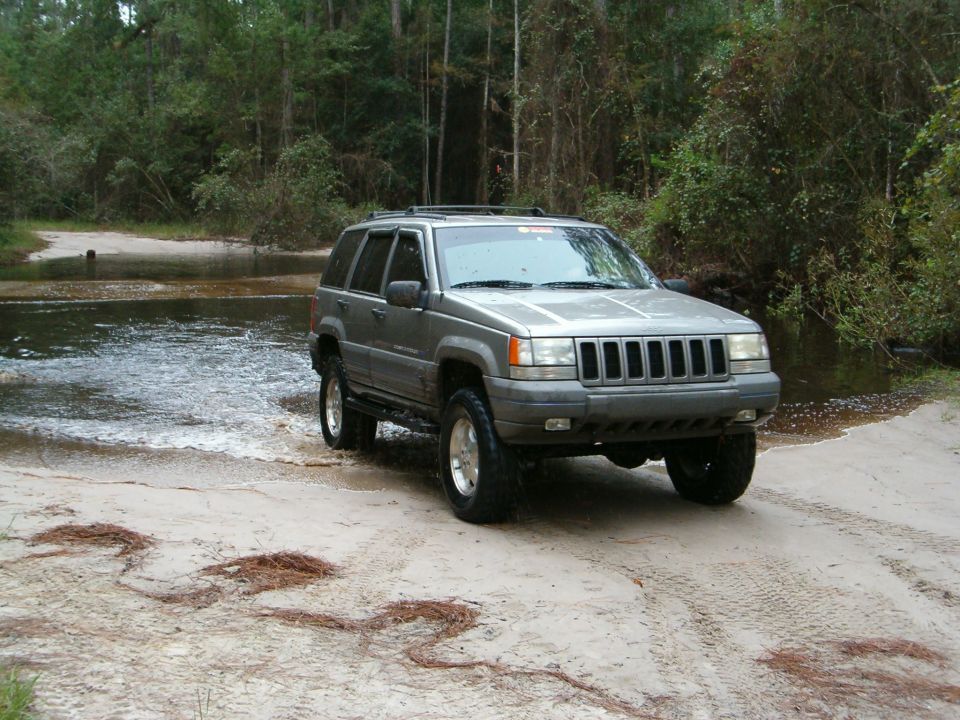 Gray Jeep Grand Cherokee ZJ exiting a flooded sandy trail crossing in pine woods