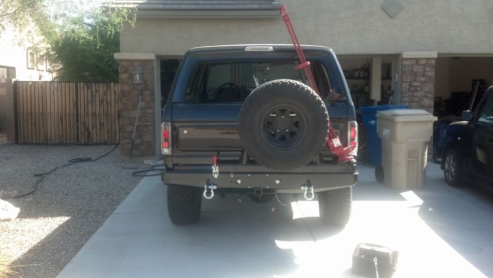 Rear view of a dark-colored Ford Bronco with a spare tire and a red Hi-Lift jack mounted on the back. It has an aftermarket bumper with tow hooks
