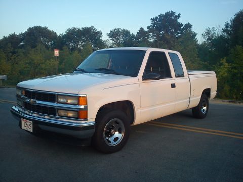 1997 Chevrolet Silverado 1500 - This white pickup truck is seen from a 3/4 front angle on a road with trees behind it.