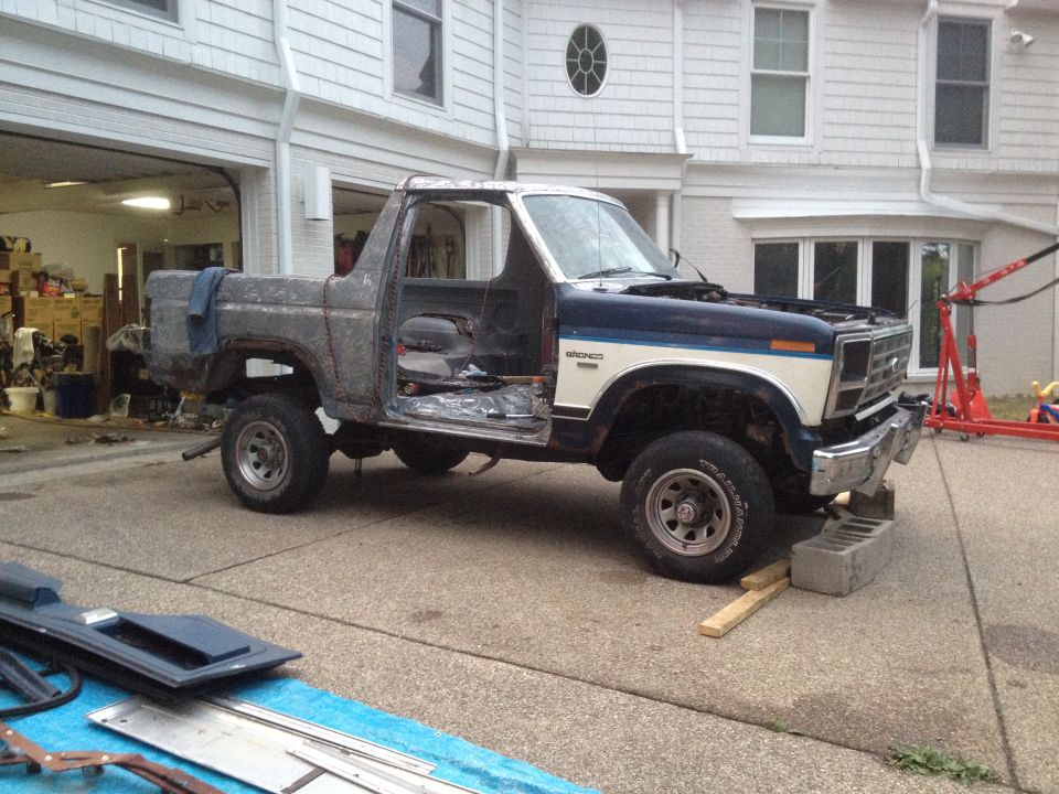 Ford Bronco with doors removed, hood up, and body in rough shape. It's parked in a driveway in front of a house