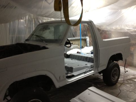 1986 Ford Bronco - White Ford Bronco undergoing restoration in a paint booth, with doors open and interior stripped.