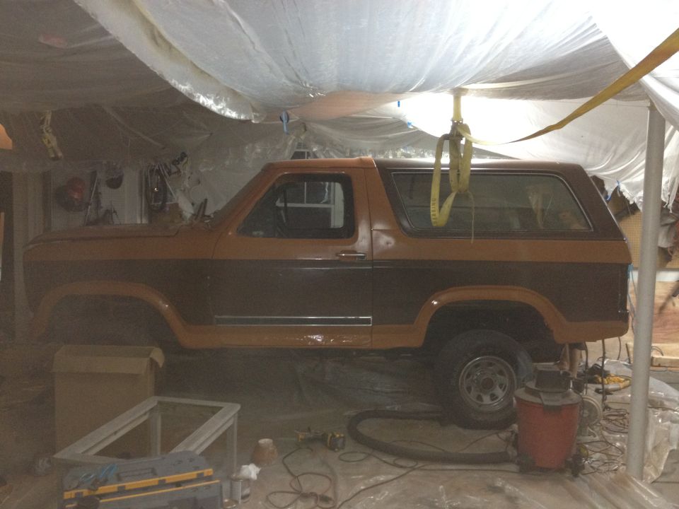 Brown and tan Ford Bronco in a garage, covered in plastic sheeting, with a yellow strap overhead