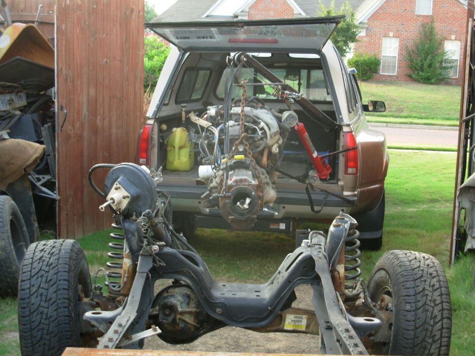 Engine hoisted in open SUV rear beside a bare vehicle frame with front axle on a trailer