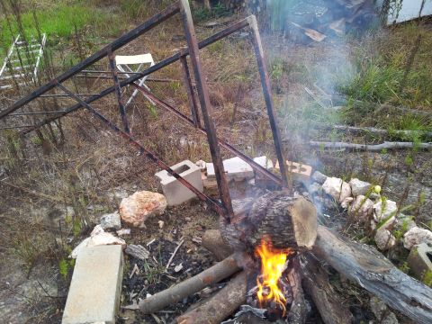 1986 Ford Bronco - A campfire burns in a rustic outdoor setting, with a metal frame overhead and a white chair nearby.