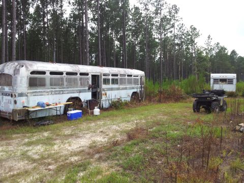 1986 Ford Bronco - An old bus and a trailer sit in a wooded area, with an ATV nearby, suggesting a remote campsite or abandoned location.