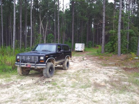 1986 Ford Bronco - A lifted black 1986 Ford Bronco sits off-road in a wooded area next to a dirt path.