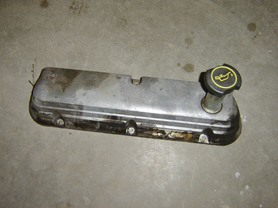 Engine valve cover with black oil cap showing oil can symbol. Part is on a concrete surface