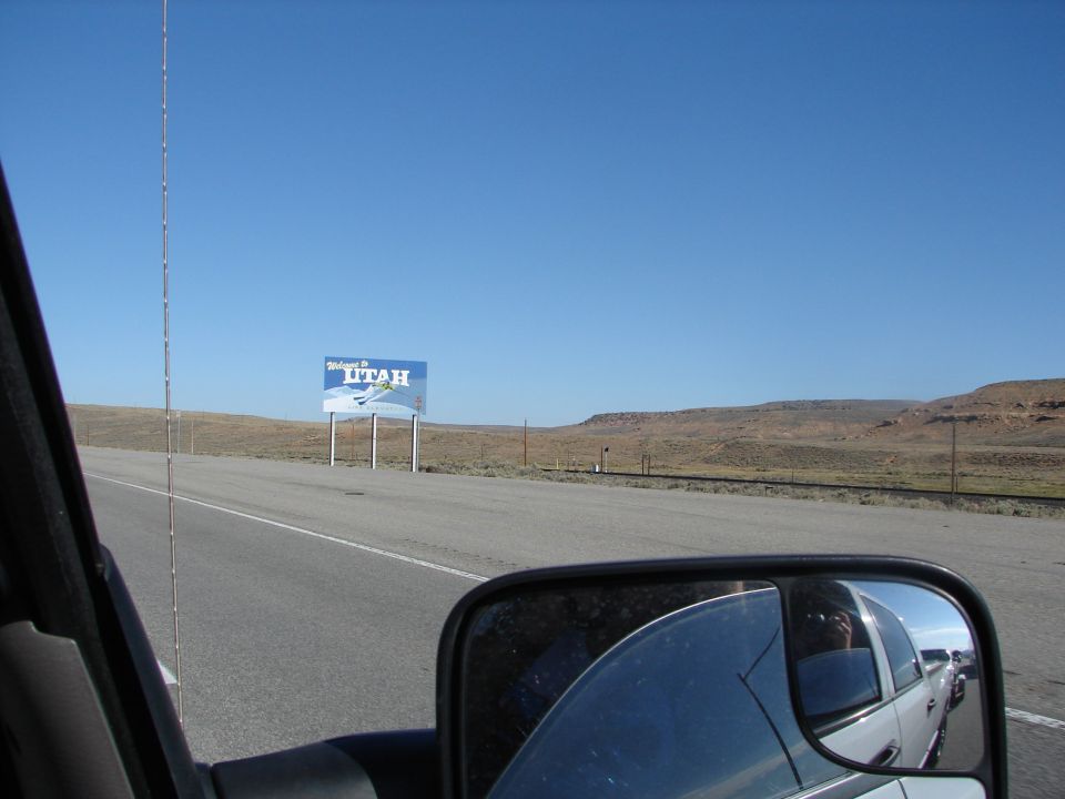 Side mirror view of white truck on highway, "Welcome to Utah" sign visible
