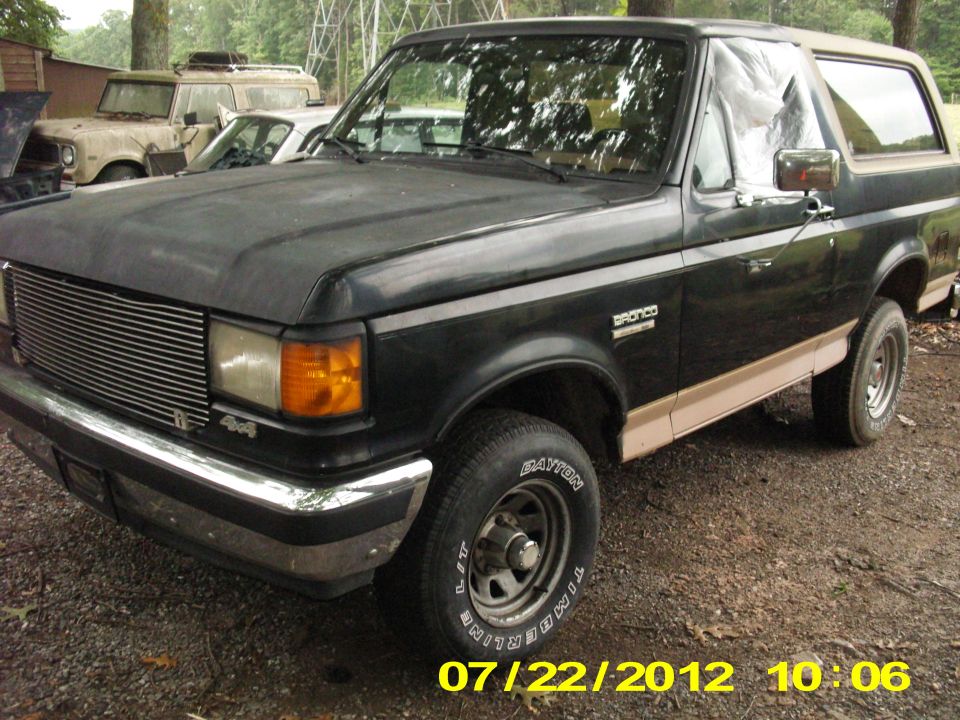 Black Ford Bronco SUV with tan lower trim, billet grille, and white-letter tires, window covered in plastic, parked on gravel.