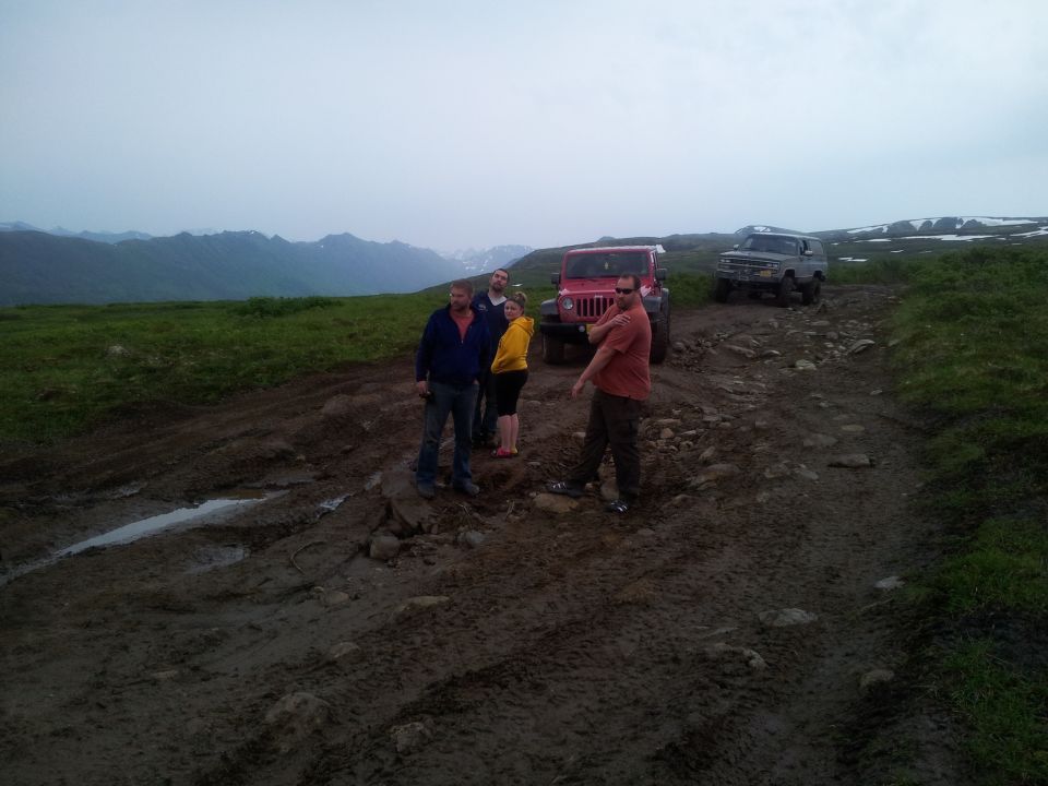 Four people stand on a muddy trail next to a red Jeep and a gray SUV, with mountains in the background