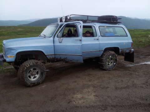 1990 Chevrolet Suburban - This light blue SUV, likely from the 1980s, is equipped for off-road conditions with large tires and chains.