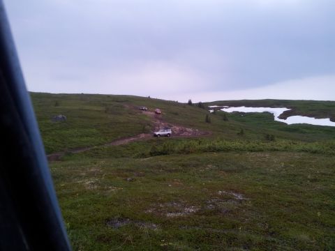 1990 Chevrolet Suburban - Several SUVs navigate a dirt track across a vast, green landscape dotted with snow.