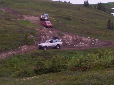 1990 Chevrolet Suburban - Three SUVs navigate a winding dirt path on a grassy incline during an off-road excursion.