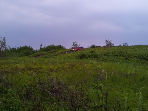1990 Chevrolet Suburban - A red SUV navigates a dirt path on a lush green hill beneath an overcast sky.