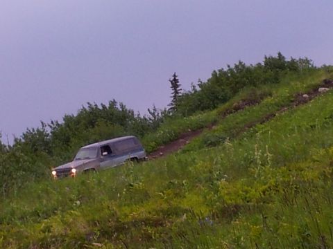 1990 Chevrolet Suburban - A 1990 Chevrolet Suburban navigates a steep, grassy incline on a dirt track under an overcast sky.