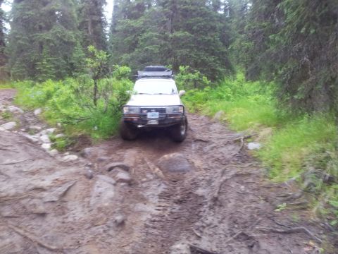 1990 Chevrolet Suburban - This white SUV navigates a challenging muddy and rocky trail through a dense forest.