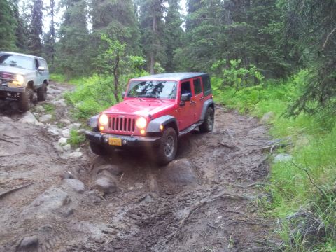1990 Chevrolet Suburban - Two off-road vehicles, a red Jeep Wrangler and a light-colored SUV, traverse a challenging muddy path.