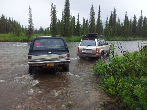 1990 Chevrolet Suburban - Two SUVs, a Chevrolet Suburban and a Toyota Land Cruiser, navigate a river in a wooded area.