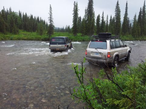 1990 Chevrolet Suburban - Two SUVs navigate a shallow river surrounded by a dense forest under an overcast sky.