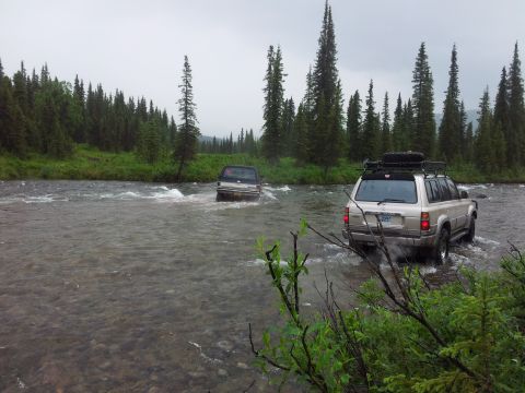 1990 Chevrolet Suburban - Two SUVs navigate a shallow river surrounded by dense forest under an overcast sky.