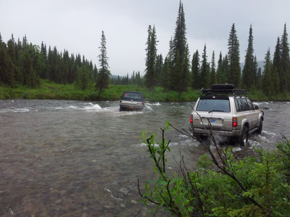 Silver SUV with roof rack and spare tire crossing a river, followed by a dark SUV