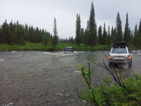 1990 Chevrolet Suburban - Two SUVs navigate a river crossing surrounded by dense forest under an overcast sky.