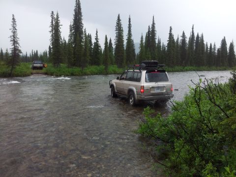 1990 Chevrolet Suburban - Two SUVs navigate a river crossing surrounded by dense pine forest.