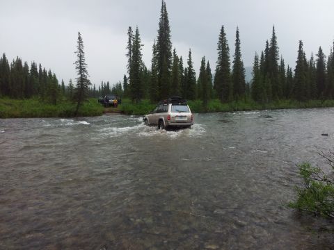 1990 Chevrolet Suburban - Two SUVs navigate a shallow river during an off-road excursion through a forested area.
