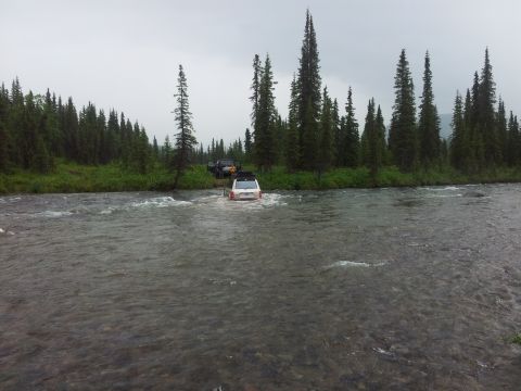 1990 Chevrolet Suburban - Two SUVs navigate a shallow river crossing amidst a dense forest under an overcast sky.