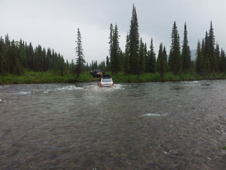 White SUV fording a river with a black SUV behind it, surrounded by trees