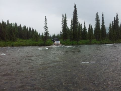 1990 Chevrolet Suburban - Two SUVs navigate a river crossing surrounded by dense evergreen forest under an overcast sky.