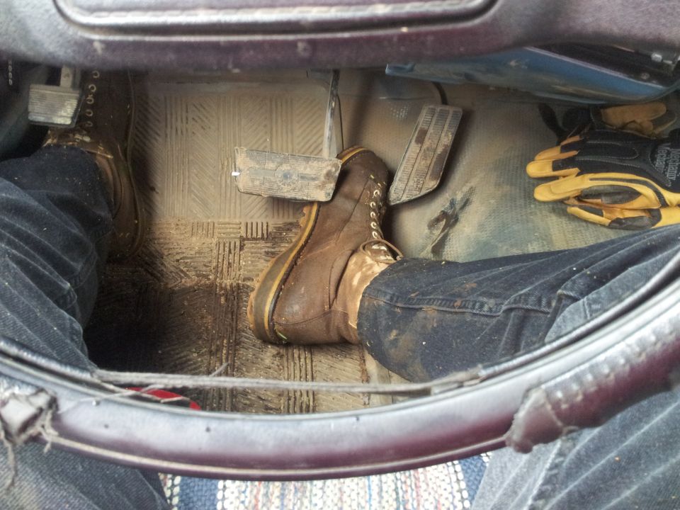 Driver's side floorboard with muddy boots on pedals and yellow gloves on the right