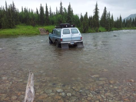 1990 Chevrolet Suburban - An older model SUV navigates a shallow river, its rear end submerged in the water.