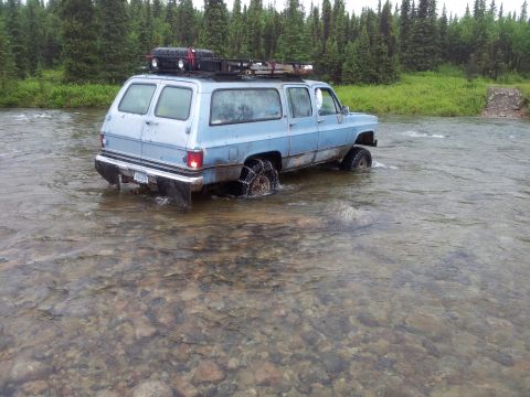 1990 Chevrolet Suburban - A light blue Chevrolet Suburban navigates a shallow river, its tires equipped with chains for traction.