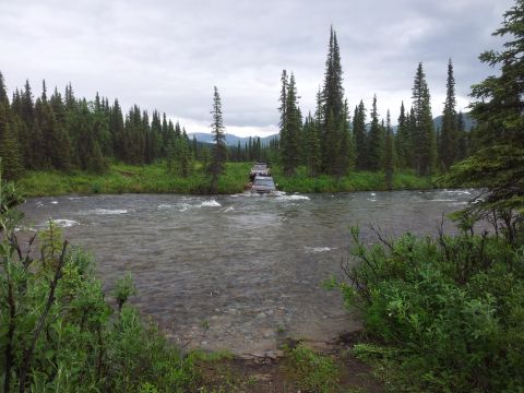 1990 Chevrolet Suburban - Several SUVs navigate a river crossing in a remote, forested area under an overcast sky.