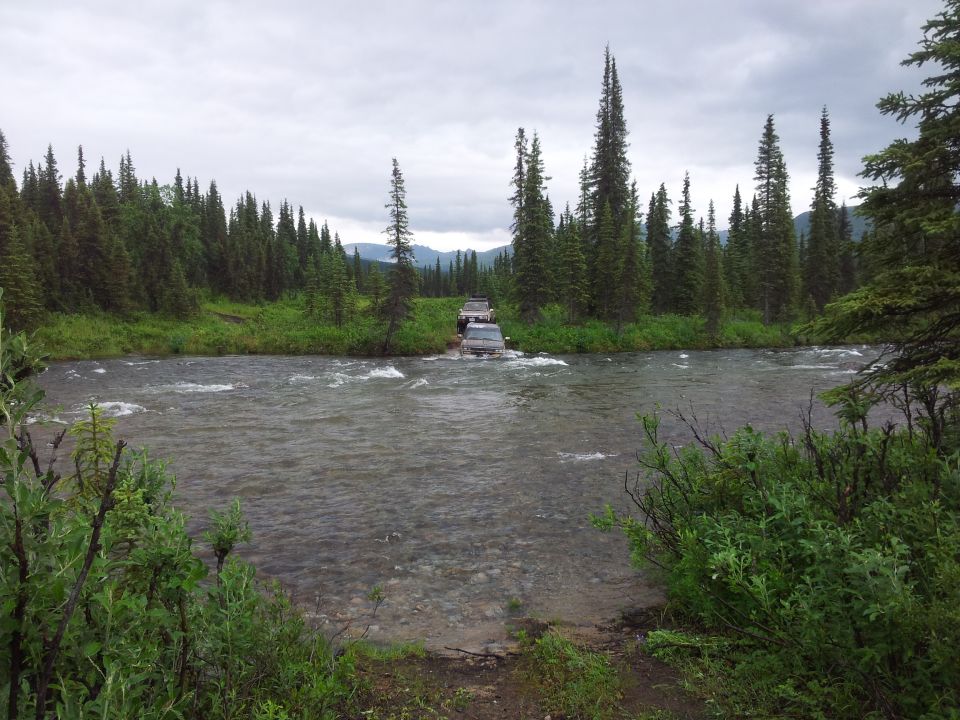 Dark SUVs crossing a shallow river with trees and mountains in the background