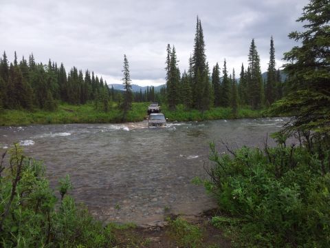 1990 Chevrolet Suburban - Two SUVs navigate a river crossing surrounded by a dense forest under an overcast sky.
