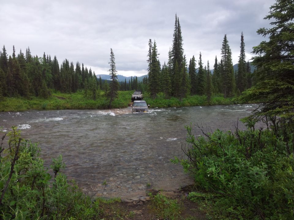 Two dark SUVs crossing a river, with dense pine forest on the banks