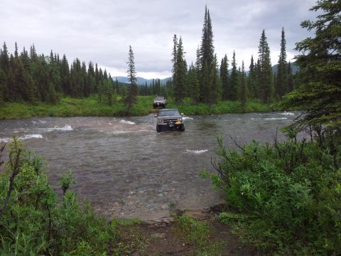 1990 Chevrolet Suburban - Two SUVs navigate a river crossing surrounded by dense pine forest under an overcast sky.