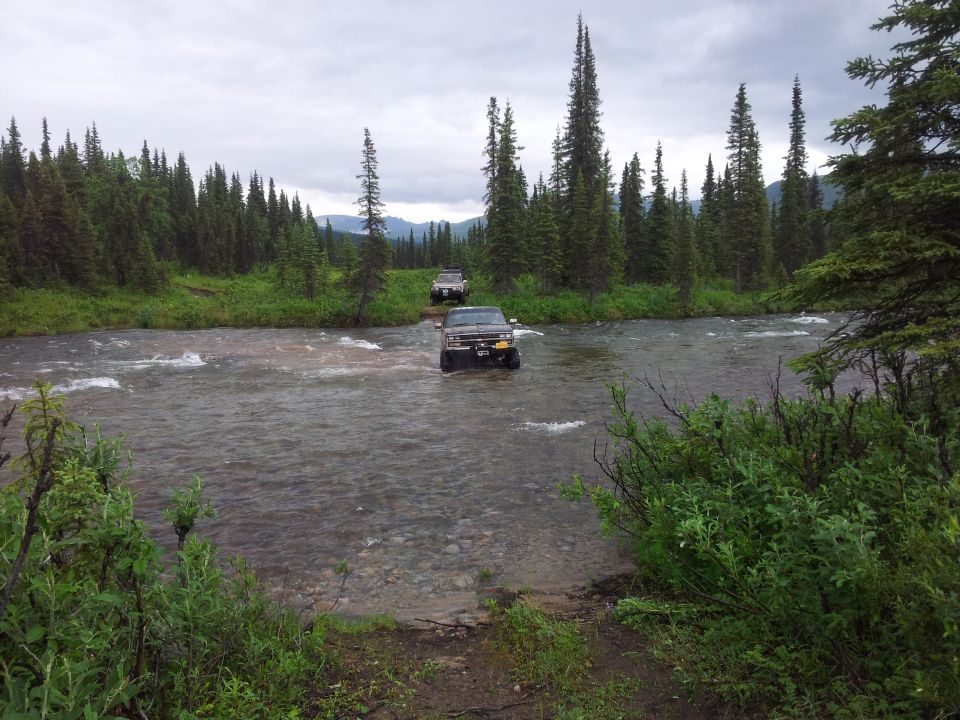 1990 Chevrolet Suburban and another SUV crossing a river in a wooded area