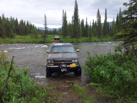 1990 Chevrolet Suburban - Two SUVs navigate a shallow river crossing surrounded by dense pine forest under an overcast sky.
