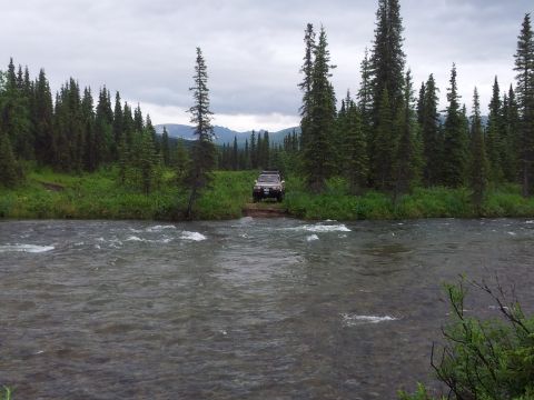 1990 Chevrolet Suburban - A dark SUV is parked on a dirt path beside a flowing river, surrounded by dense pine forest under a cloudy sky.